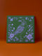 Square ceramic coaster featuring intricate lavender illustration of a raven surrounded by botanical elements on forest green background, with ornate border design and cork backing.